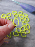 Yellow 3D printed stitch markersheld by a hand on a wooden surface with a purple tool in the background.