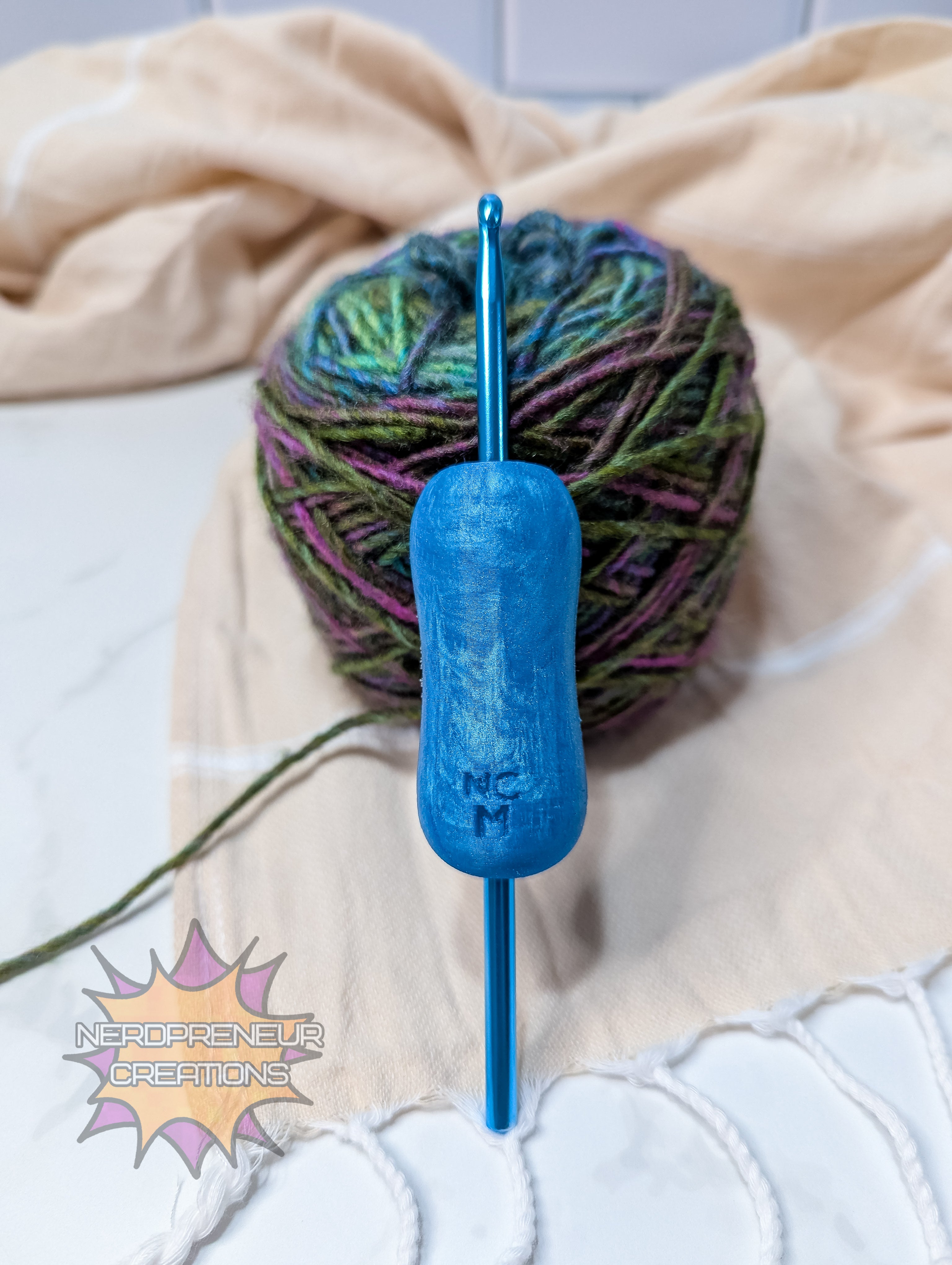 Blue silicone crochet grip and multicolored yarn on a beige fabric background with 'Nerdpreneur Creations' logo.