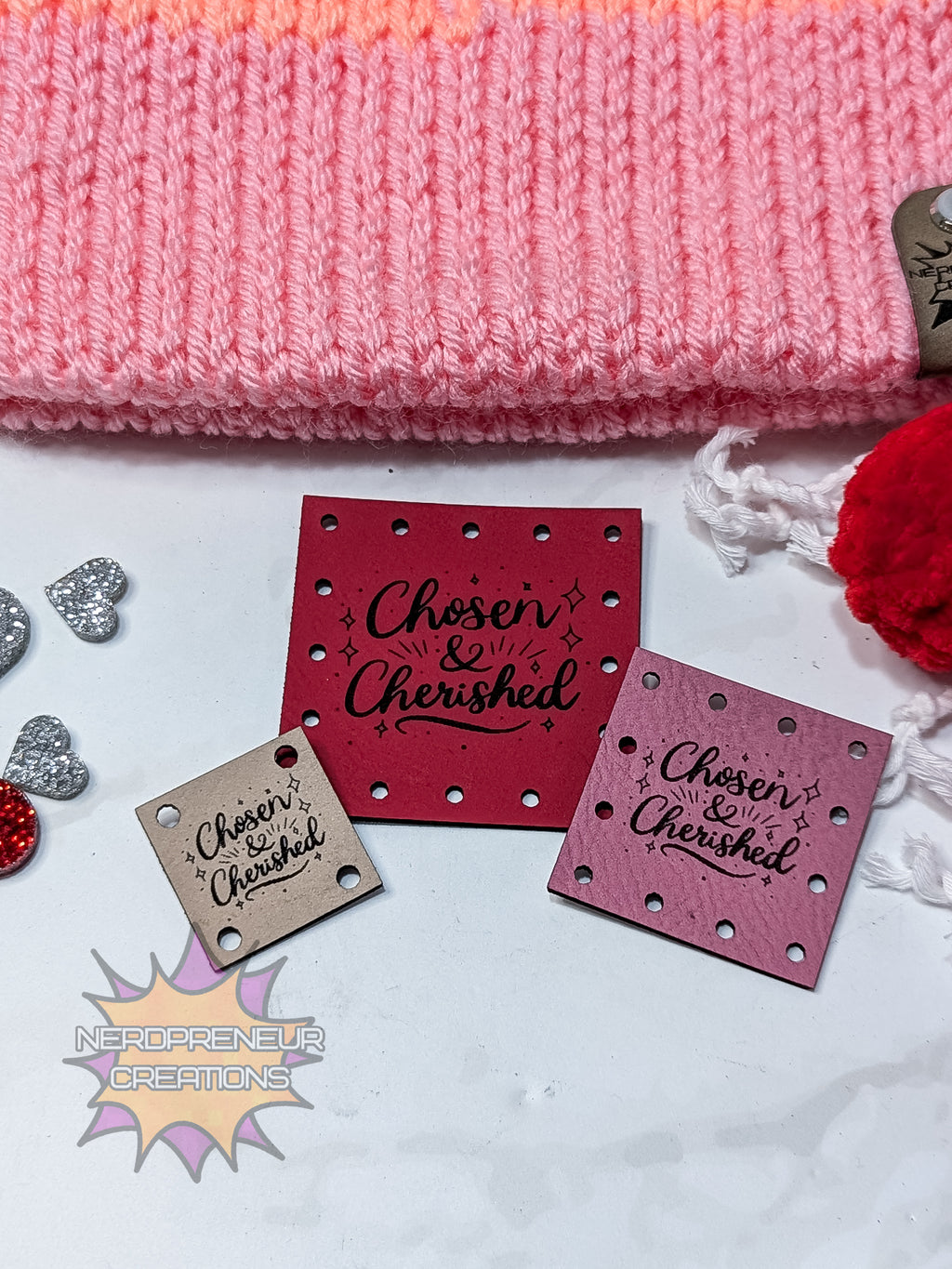 Decorative faux leather patches with 'Chosen & Cherished' text on a white surface with pink fabric and red pom-pom.