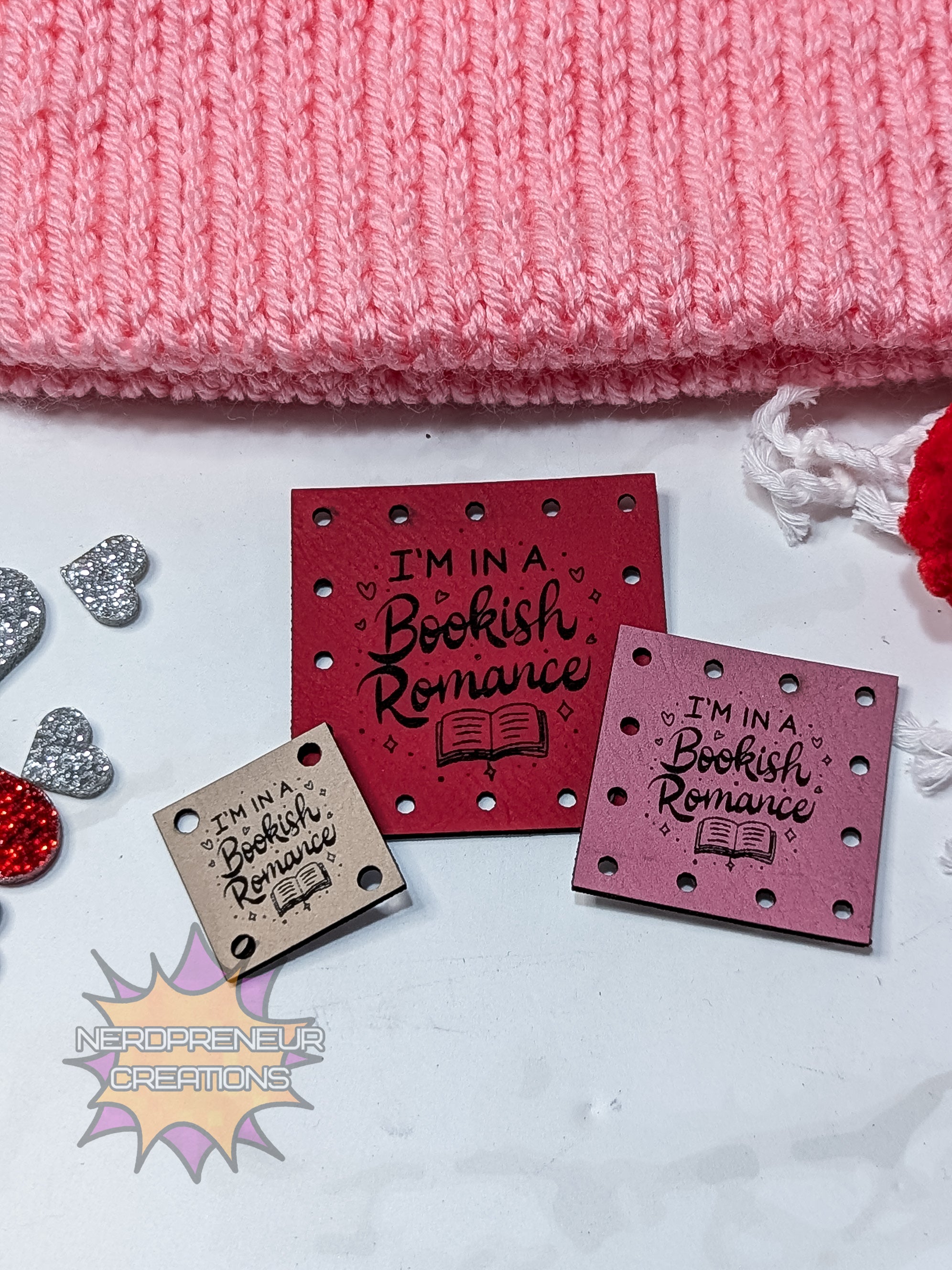 Red square faux leather patches with 'I'm in a Bookish Romance' text on a pink knitted fabric background.