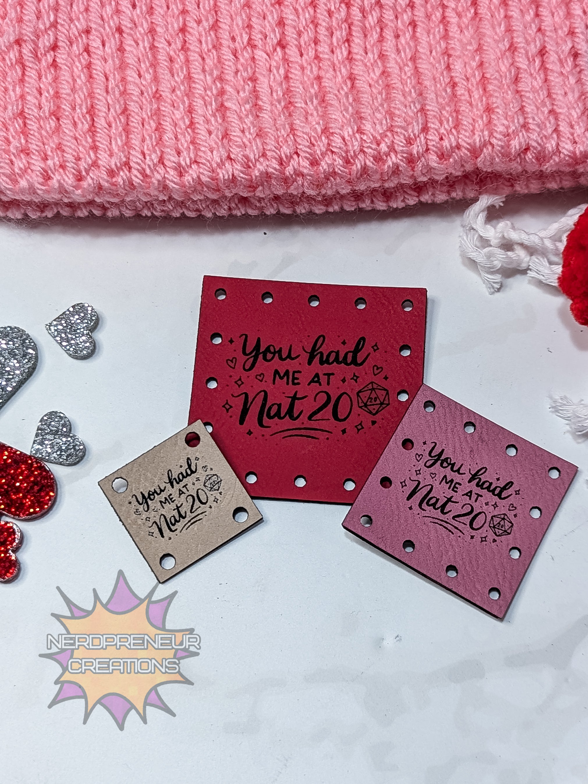 Three square metal tags with text 'You had me at Nat 20' on a pink knitted fabric background.