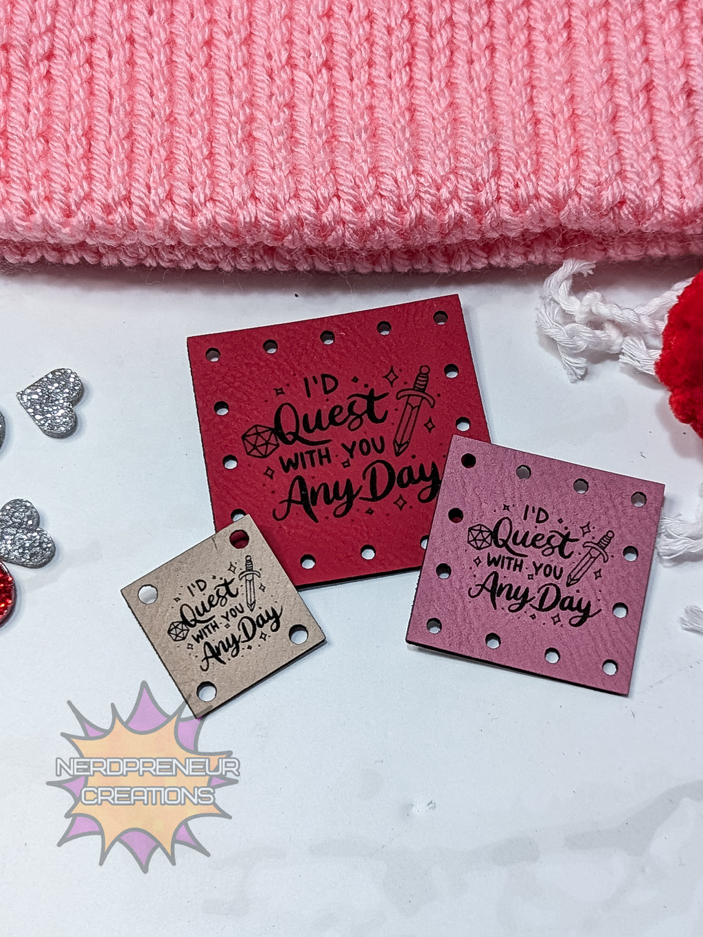 Two red faux leather patches with 'I'd Quest With You Any Day' text on a white surface with pink fabric in the background.
